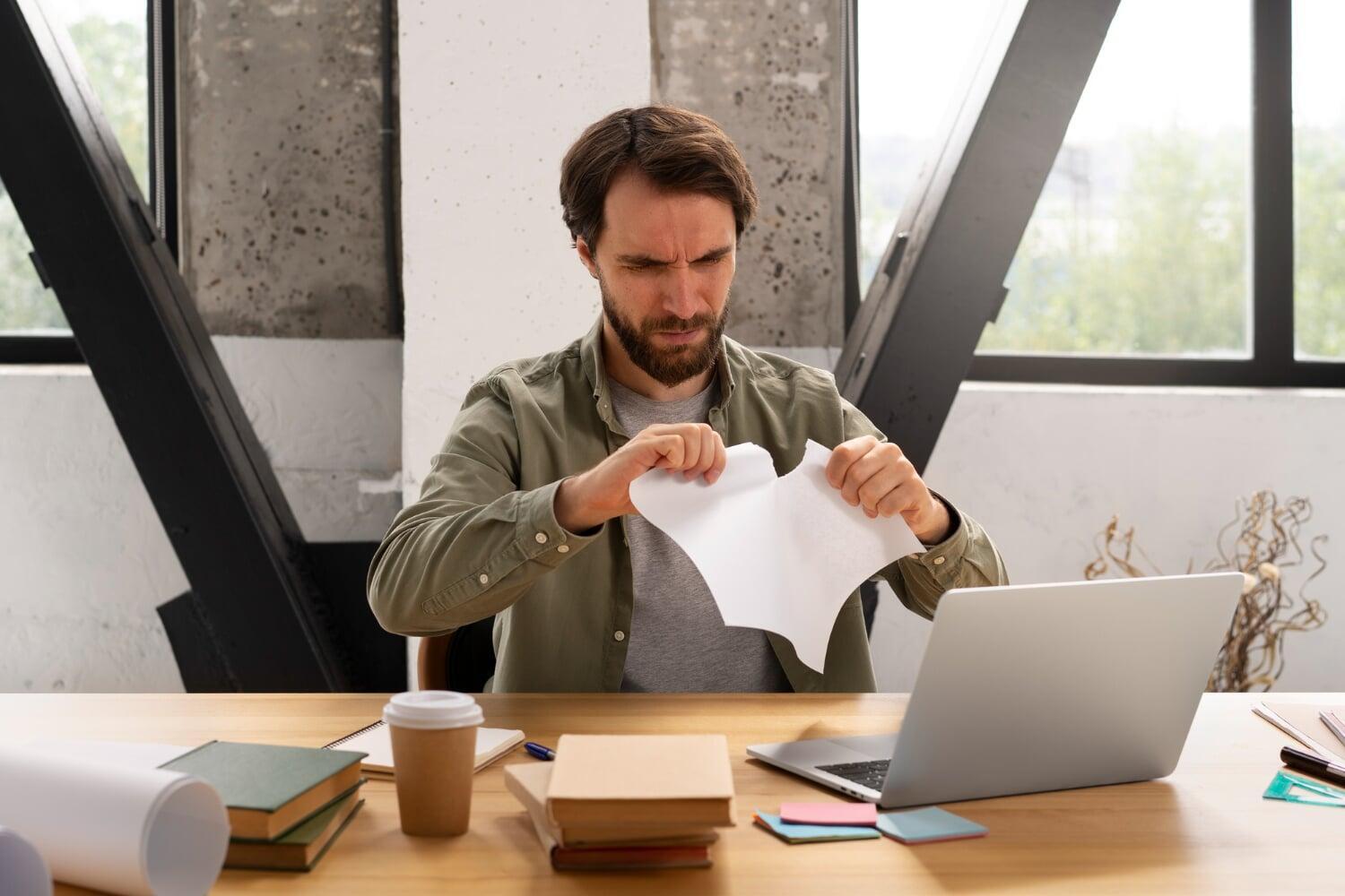 Homem rasga papel com expressão de frustração em frente a notebook
