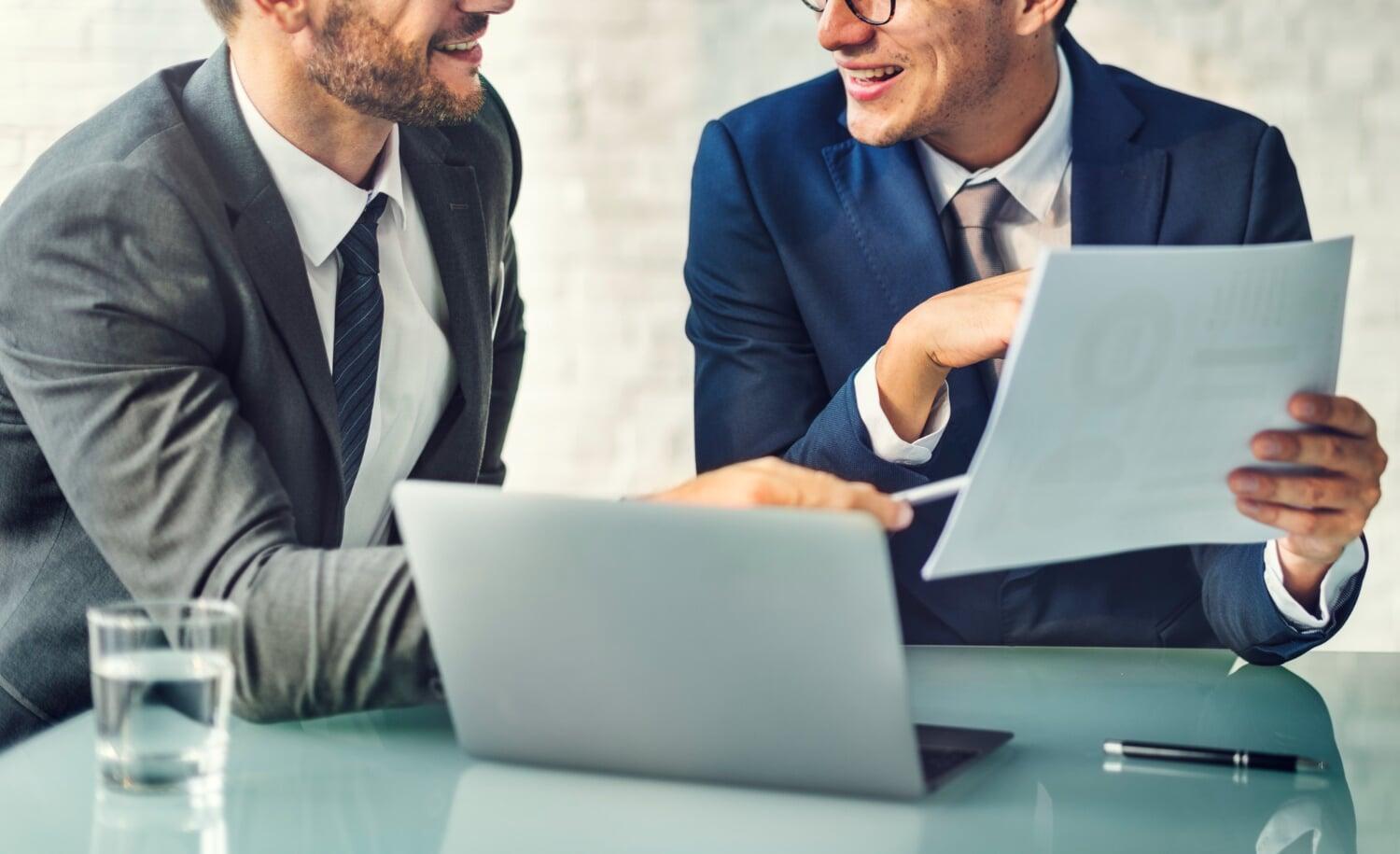 dois homens de terno analisando papel com laptop sobre a mesa