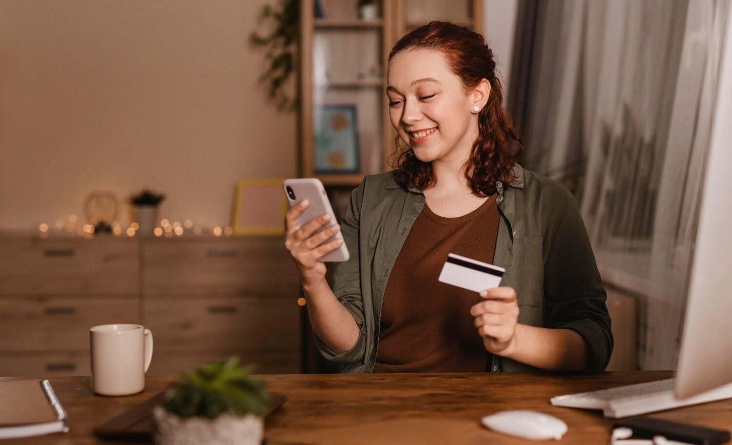 Mulher sorrindo com celular e cartão de crédito na mão, realizando pagamento online
