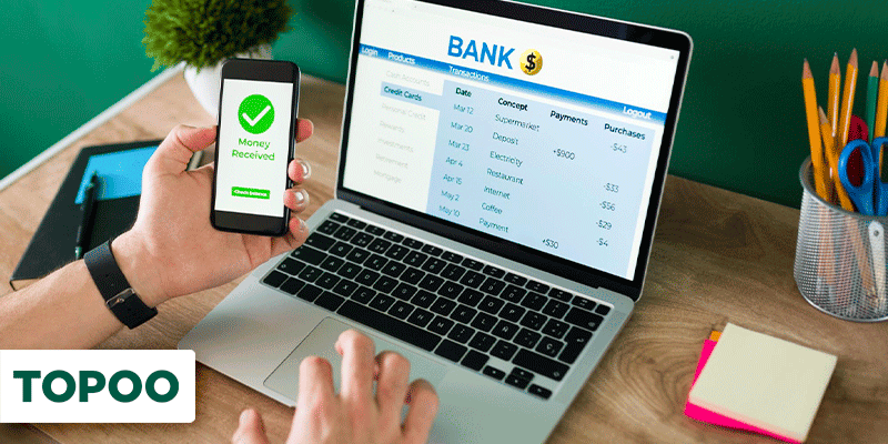 Pessoa usando celular com aplicativo bancário e notebook aberto em tela de transações financeiras