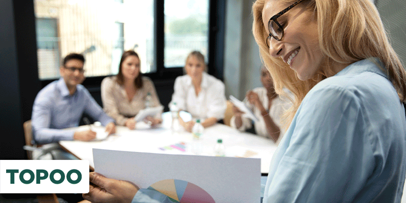 Apresentadora loira mostrando gráfico colorido em reunião com equipe ao fundo