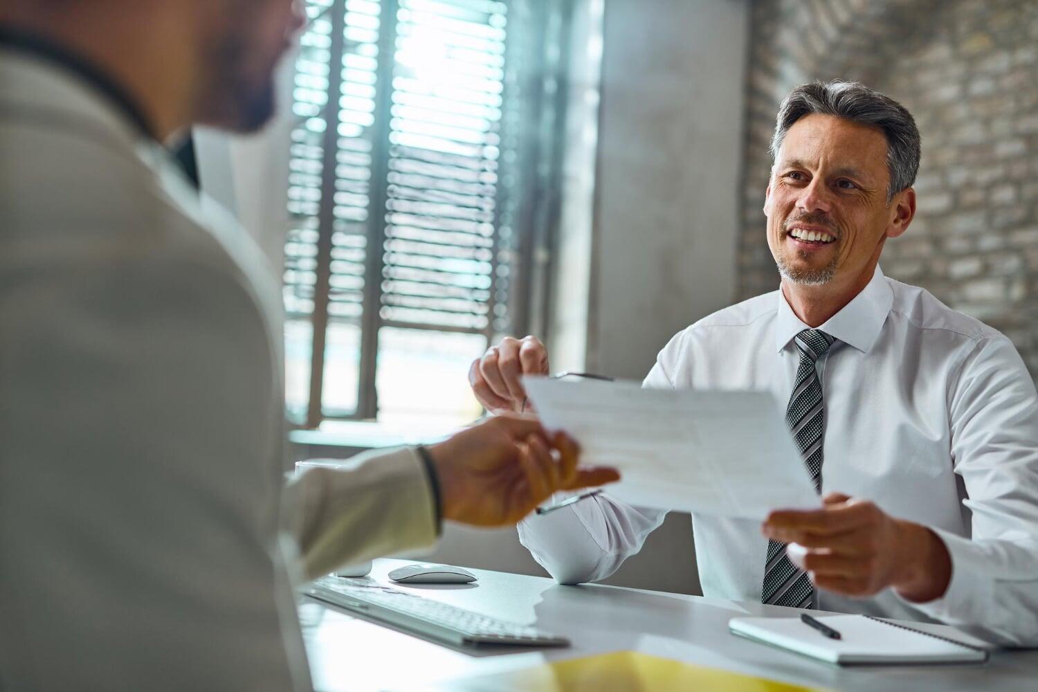 Homem sorridente segurando documento em conversa com outra pessoa em ambiente corporativo