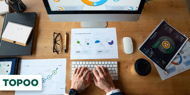 Profissional analisando gráficos financeiros em tela de computador e documentos sobre a mesa
